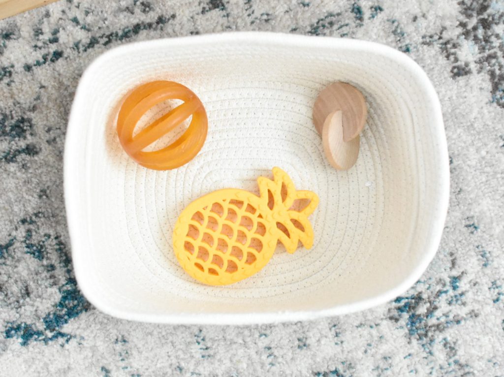 The image shows a white basket with a Calmies natural teether, interlocking discs, and a pineapple teether in it.