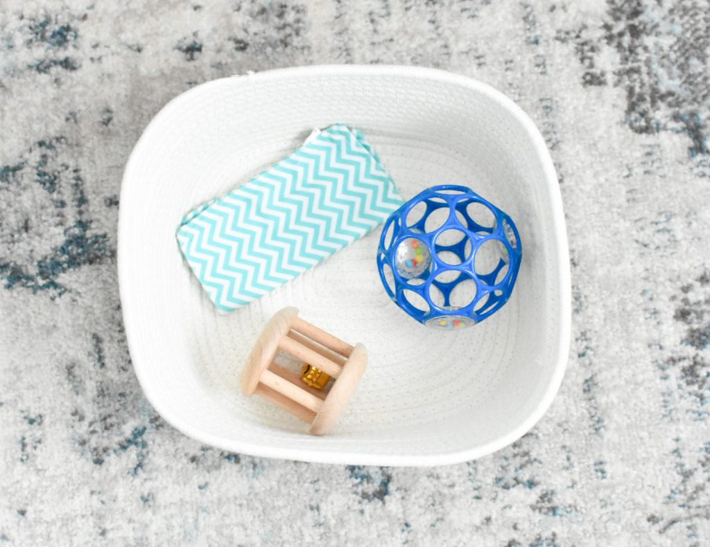 The image shows a white basket with baby paper, a blue Oball, and a wooden bell rattle in it.